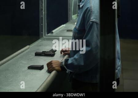 Mann in Jeansjacke, der bei der Schusswaffenkontrolle auf dem Schießstand in der Halle war. Schusswaffe genau beobachten, Hand auf der Theke ruhen, eine weitere Waffe und ein Magazin in der Nähe auf der Theke platziert Stockfoto