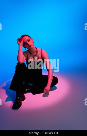 Eine stilvolle junge Frau sitzt anmutig in rosa und blauer Beleuchtung und zeigt moderne Mode. Stockfoto