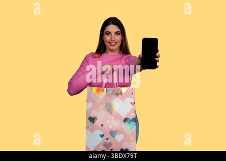 Lächelnde Frau, die einen leeren Smartphone-Bildschirm zeigt, während sie eine Einkaufstasche mit Herzmuster vor gelbem Hintergrund hält Stockfoto