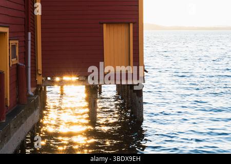 Traditionelle rote norwegische Fischerhütten auf Stelzen am Fjord, beleuchtet von der untergehenden Sonne mit Reflexen auf dem Wasser. Stockfoto
