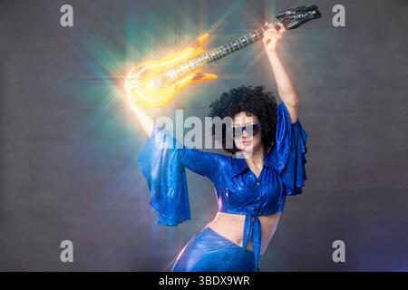 Mädchen-Comic-Popstar posiert in schwarzer Perücke mit Spielzeuggitarre auf blauem Hintergrund. Rock-Stil der 70er Jahre Stockfoto