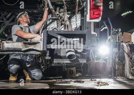 Ein Mechaniker arbeitet bei Tageslicht in einer Werkstatt, die mit Werkzeugen und Drähten gefüllt ist, mit Sorgfalt am Fahrwerk eines Fahrzeugs. Die Umgebung ist sehr beschäftigt und beschäftigt Stockfoto
