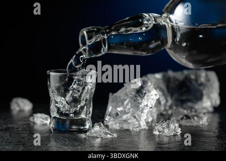 Kristallklarer Wodka oder Gin wird in ein Glas gegossen. Transparentes alkoholisches Getränk mit Eisstücken auf schwarzem Hintergrund. Stockfoto