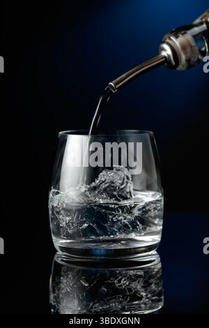 Kristallklarer Wodka oder Gin wird über Eis in ein Glas gegossen. Wodka mit Eis auf schwarzem reflektierendem Hintergrund. Stockfoto