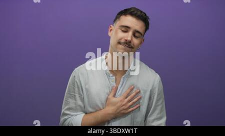 Junger Mann, der vor einer leuchtenden lila Wand Emotionen ausdrückt, ein lässiges Hemd trägt und seine Brust berührt, wobei er die hispanic-Abstieg und einen IS hervorhebt Stockfoto