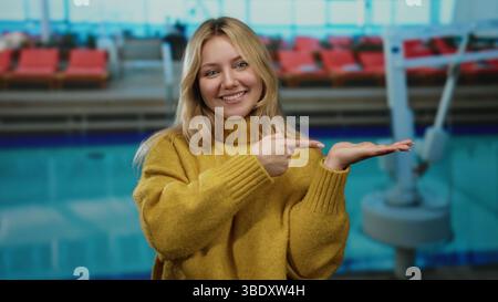 Frau lächelte am Pool im Resort und zeigte auf offene Hand mit gelbem Pullover entspannte Atmosphäre im Freien. Stockfoto