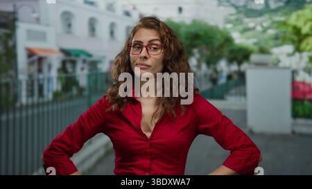 Selbstbewusste Frau in rotem Hemd steht mit Armen akimbo auf einer Stadtstraße und vermittelt Entschlossenheit und Selbstbewusstsein in einer urbanen Outdoor-Umgebung. Stockfoto