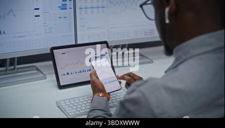 Finanzanalyst überprüft Echtzeitaktien, Börsenmarkttafeln auf Smartphone und digitale Displays mit der Online Trading App. Afroamerikanischer Geschäftsmann arbeitet im Investment Bank Office. Nahaufnahme. Stockfoto