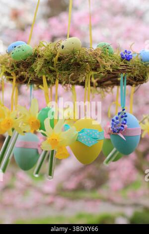 Osterkranz. Einfach zu badizierendes Kranzprojekt mit Moos, Miniatur-Narzissen und Osterei, aufgehängt auf Gartenzweig mit gelbem Band. UK Stockfoto