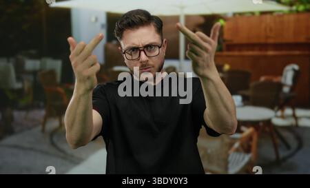 Junger Mann mit Bart, der auf der Terrasse des Stadtcafés unhöfliche Gesten macht, umgeben von einem Restaurant im Freien mit Tischen und Stühlen. Stockfoto