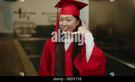 Frau in roter Abschlusskleidung steht lächelnd in einer Küche und macht eine Handygeste, die Leistung in einer Inneneinrichtung verkörpert. Stockfoto