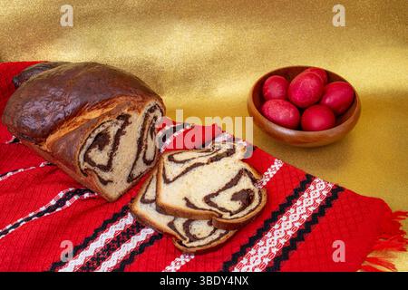 Rumänisches Süßbrot „Cozonac“, gefüllt mit Walnüssen und Schokolade, auf einem roten traditionellen Handtuch, ostereier auf goldenem Hintergrund Stockfoto