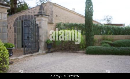 Verschwommener Blick auf einen mediterranen Garten mit einer alten Steinmauer und einem schmiedeeisernen Tor, umgeben von üppigem Grün, schaffen eine ruhige und zeitliche Atmosphäre Stockfoto