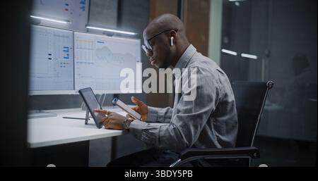 Afroamerikanischer Finanzanalyst, der Aktien in Echtzeit analysiert, Börsenkarten auf einem digitalen Tablet, Smartphone und Computer mit Multi-Monitor-Workstation ausgibt. Geschäftsmann im Investment Bank Office. Stockfoto