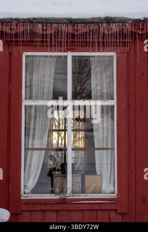 Traditionelles rotes Holzhaus. Fenster mit weißem, dünnem Rahmen und einem Vorhang, Blick auf das gegenüberliegende Fenster. Eiszapfen am Dachrand mit Schnee. Hars Stockfoto