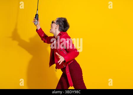 Energiegeladene Seniorin, die leidenschaftlich singt, während sie ein Mikrofon in stilvoller Mode vor einem leuchtend gelben Hintergrund hält Stockfoto