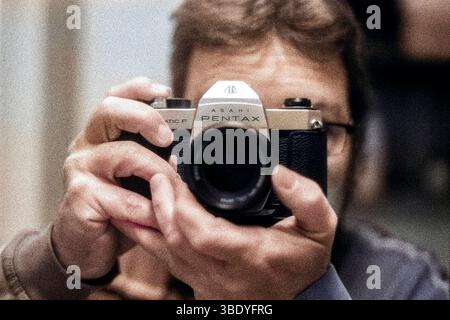 Selfie/Selbstporträt eines erwachsenen Mannes, der eine Brille trägt und eine 50 Jahre alte analoge Pentax Film- und Fotokamera verwendet. Aufgenommen mit analogem Schwarzweißfilm, farbig mit PS Neural Filtern. Tilburg, Niederlande. Stockfoto