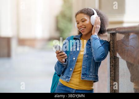 Junge schwarze Studentin, die Musik über Kopfhörer hört und ihr Smartphone überprüft Stockfoto