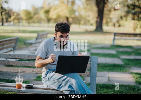 Fröhlicher Mann, der Erfolg feiert, während er im Park draußen mit einem Laptop unterwegs ist Stockfoto