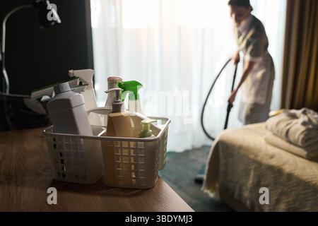 Person saugt den Raum ab, während verschiedene Reinigungsmittel im Vordergrund auf dem Tisch sitzen. Helles Licht aus dem Fenster beleuchtet die Szene Stockfoto