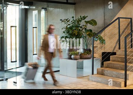 Verschwommenes Bild eines Mannes in Geschäftskleidung, der einen Koffer trägt, während er durch die geräumige, moderne Bürolobby mit markanten Pflanzen und einer Metalltreppe im Hintergrund geht Stockfoto