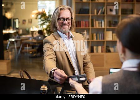 Porträt eines älteren Mannes, der lächelt, während er seine Kreditkarte in einer gemütlichen Bibliothek überreicht, und mit einem Bibliotheksmitarbeiter interagiert, der im Vordergrund nicht scharf ist Stockfoto
