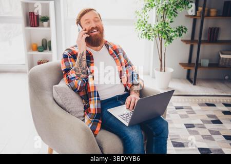 Mann mittleren Alters mit Bart, der tagsüber im gemütlichen Wohnzimmerinnenraum telefoniert, während er einen Laptop benutzt Stockfoto