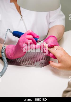 Nahaufnahme eines professionellen Nageltechnikers, der Nägel mit einem elektrischen Feilwerkzeug formt, mit rosa Handschuhen in einem sauberen Salon. Stockfoto