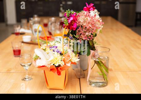 Zwei leuchtende Blumensträuße in Vasen und eine Geschenkbox auf einem Holztisch, umgeben von Getränken und Gläsern. Stockfoto
