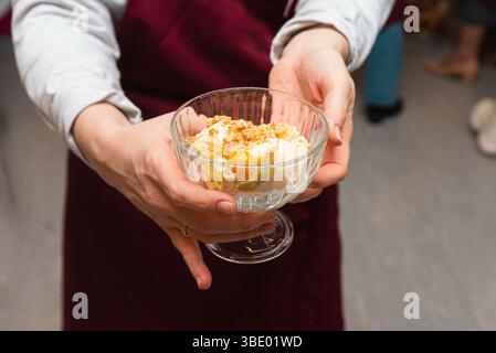 Person hält ein schickes Dessert mit Streuseln und Physalis in einer klaren Glasschale, die zum Servieren oder Genießen bereit ist. Stockfoto