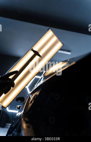 Bei der lacklosen Dellen-Reparatur bei PDR werden Dellen unter Lichtreflexion für Präzision geschlagen. Der Techniker verwendet PDR-Werkzeuge, um Dellen von der Motorhaube zu entfernen Stockfoto