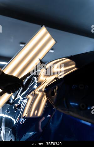 Bei der lacklosen Dellen-Reparatur bei PDR werden Dellen unter Lichtreflexion für Präzision geschlagen. Der Techniker verwendet PDR-Werkzeuge, um Dellen von der Motorhaube zu entfernen Stockfoto