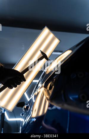 Bei der lacklosen Dellen-Reparatur bei PDR werden Dellen unter Lichtreflexion für Präzision geschlagen. Der Techniker verwendet PDR-Werkzeuge, um Dellen von der Motorhaube zu entfernen Stockfoto