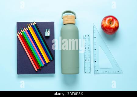 Ein Notizbuch, bunte Bleistifte, eine Flasche, ein Apfel und Lineale auf hellblauem Hintergrund. Back-to-School-Konzept. Stockfoto