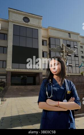 Eine sanfte junge Indianerin in medizinischer Kleidung mit Stethoskop steht vor einem Gebäude mit Säulen und großen Fenstern. Ein medizinisches Symbol im Stockfoto