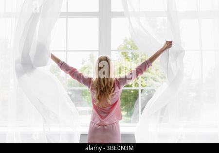 Junge hübsche blonde Frau in rosa Schlafanzug wachte auf, Blick auf große Panoramafenster im Schlafzimmer Stockfoto