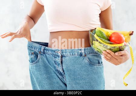 Beschnittene Ansicht der schwarzen Frau mit Schale von Früchten, Gemüse und Maßband tragen übergroße Jeans auf hellem Hintergrund Stockfoto