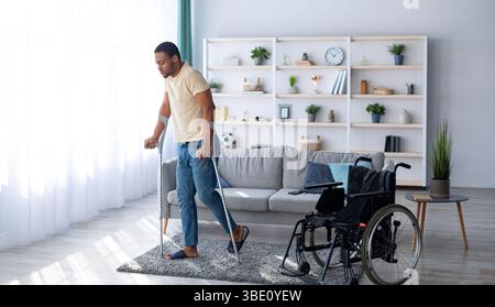 Rehabilitationskonzept. Junger schwarzer Kerl, der mit Krücken läuft, nachdem er zu Hause einen Rollstuhl benutzt hat, Panorama Stockfoto