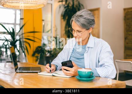 Reife Frau, die freiberuflich von Café mit Laptop, Kaffee und Smartphone arbeitet und Lifestyle ausgleicht Stockfoto