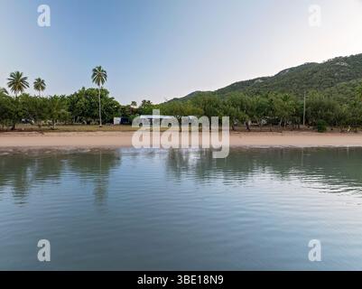 Tropische Küstenszene mit immer noch Reflexionen und sandiger Küste in der Nähe von bewaldeten Hügeln. Stockfoto