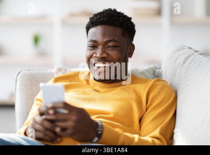Lächelnder afroamerikanischer Mann, der mit dem Handy auf der Couch liegt Stockfoto