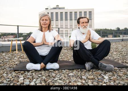 Älteres Paar übt achtsames Yoga auf dem städtischen Dach in ruhiger Meditationsposition Stockfoto