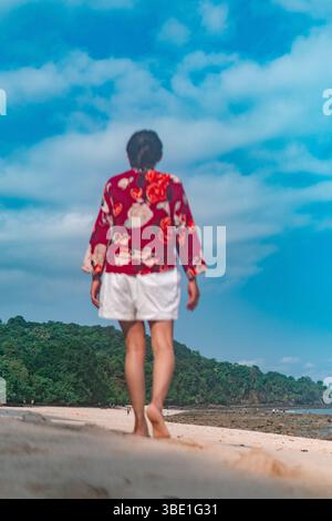 Eine Frau in einem roten Blumenhemd und weißen Shorts läuft barfuß an einem tropischen Strand. Der Himmel ist blau und grüne Hügel säumen den Hintergrund. Stockfoto