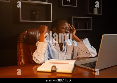 Eine junge Geschäftsfrau hat schwere Kopfschmerzen, massiert ihre Schläfen und schließt die Augen, sitzt mit Laptop und Notebook an ihrem Schreibtisch, arbeitet spät in der Nacht in ihrem Büro Stockfoto