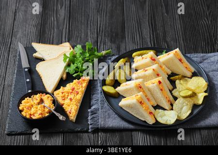 pimento-Käse-Dreieck-Sandwiches mit Gurken und Kartoffelchips auf einem Teller auf schwarzem Holztisch, horizontaler Blick von oben, amerikanische Küche, keine Stockfoto