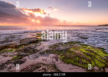 Spektakulärer Sonnenuntergang über Rocky Beach und Illa del Aire Leuchtturm in Punta Prima, Menorca. Pulsierender Himmel, Meereswellen, zerklüftete Küste, mediterranes Be Stockfoto