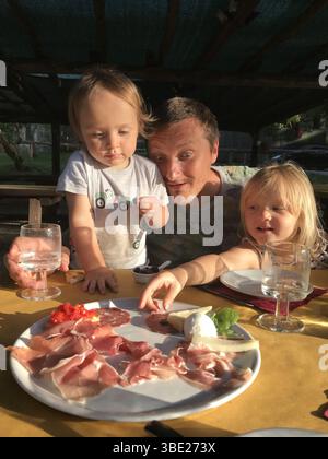 Glückliche Familie genießt einen Teller italienische Antipasti während eines Picknicks im Freien in der Sonne. Zwei Söhne und ein Vater schauen mit Begeisterung auf die Vielfalt von Stockfoto