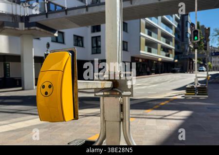 Ein hellgelber Fußgängerknopf ist an einem Ständer in einer modernen städtischen Umgebung montiert. In der Nähe ist die Straße ruhig, mit Gebäuden und einer Straße Stockfoto