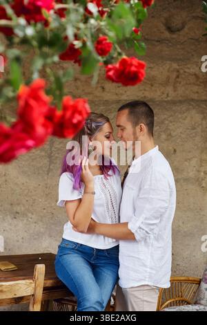 Ein Paar teilt einen intimen Moment in einem malerischen Außenbereich, der mit leuchtend roten Rosen geschmückt ist. Sie sind in liebevoller Umarmung verwickelt, lässig gekleidet Stockfoto
