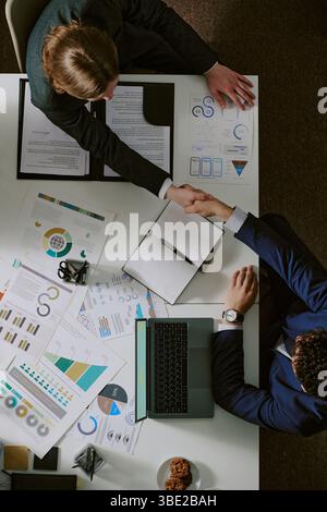 Draufsicht auf Geschäftsleute, die die Hände über den Schreibtisch schütteln, mit Diagrammen, Diagrammen und einem Notebook Stockfoto